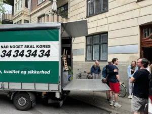 Flyttevogn laster møbler ved opgang; kundens eget flyttehold bærer indbo op ad trapper — vi leverer transport og indlastning.