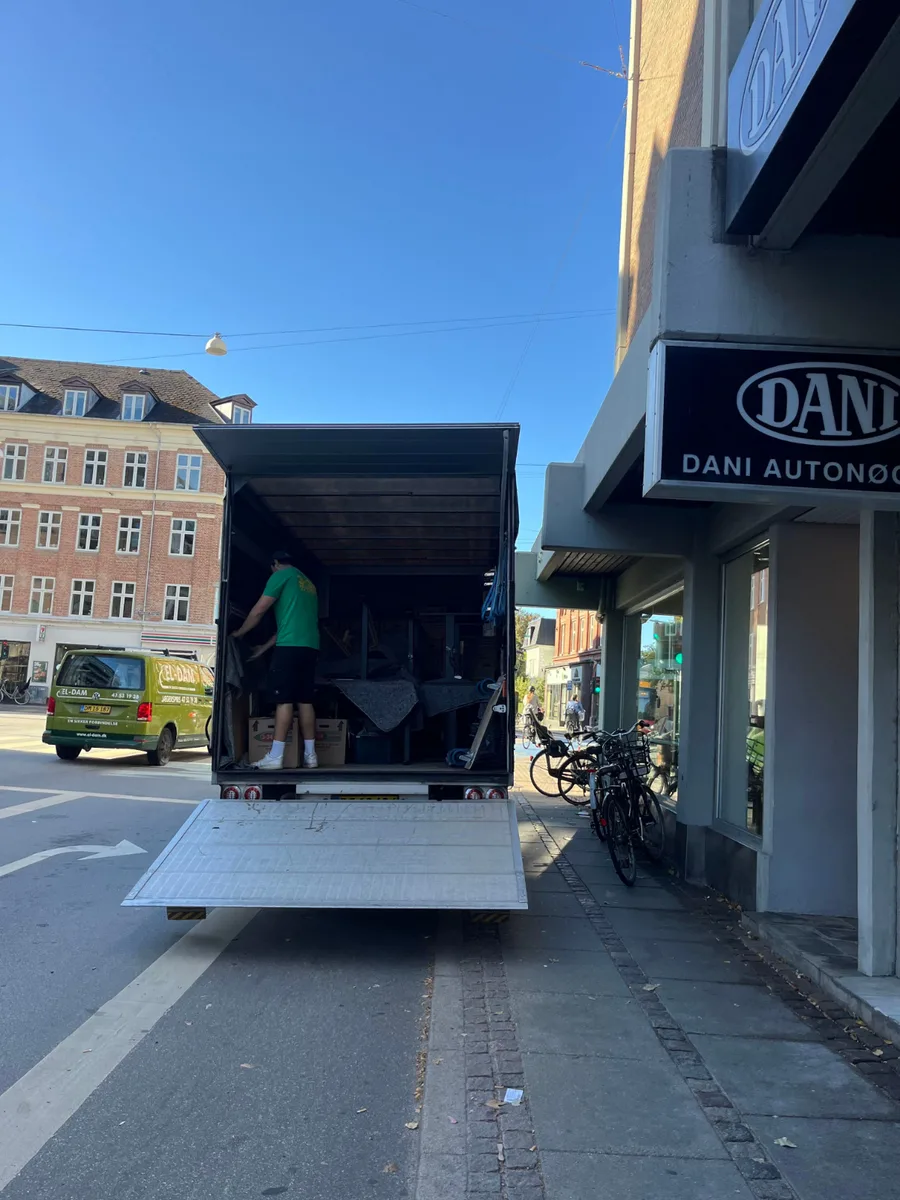 En flyttevogn med sænket rampe holder ved fortovskanten, mens en flyttemand arbejder inde i ladet.