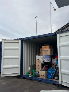 En person ligger ved åbningen til en åben container fyldt med flyttekasser, plastkasser, plastposer og en støvsuger.