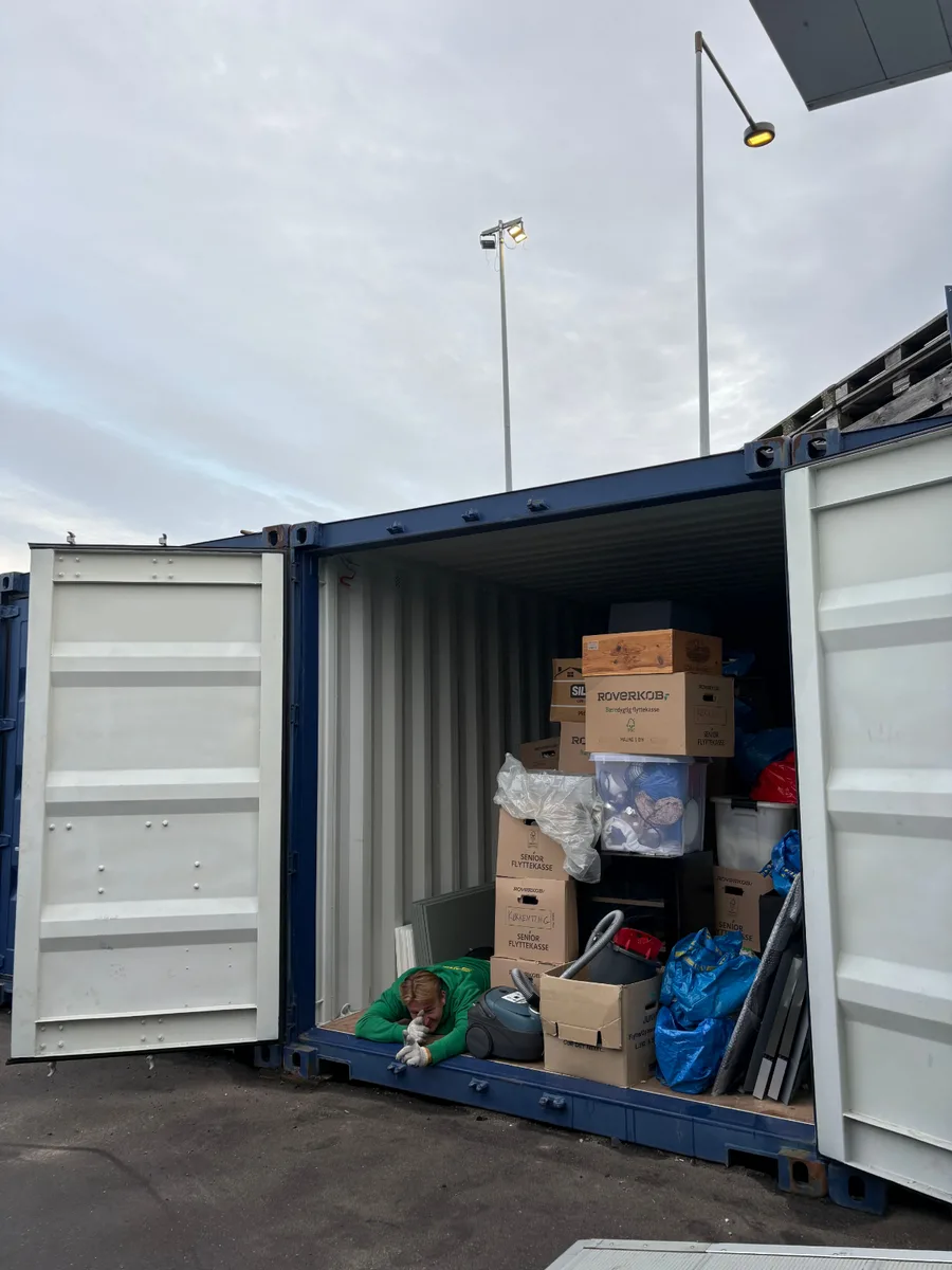 En person ligger ved åbningen til en åben container fyldt med flyttekasser, plastkasser, plastposer og en støvsuger.
