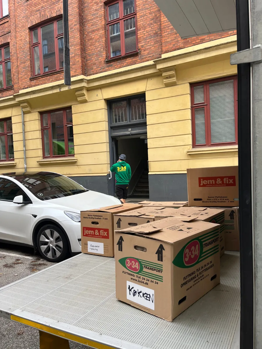 Flyttekasser står på en flyttevogn ved en opgang, mens en person står ved døren.