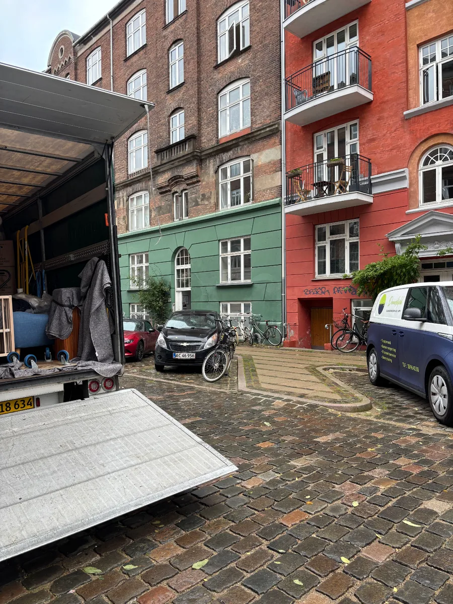 En åben flyttevogn med møbler indpakket i flyttetæpper står i en brolagt gård mellem boligbygninger.