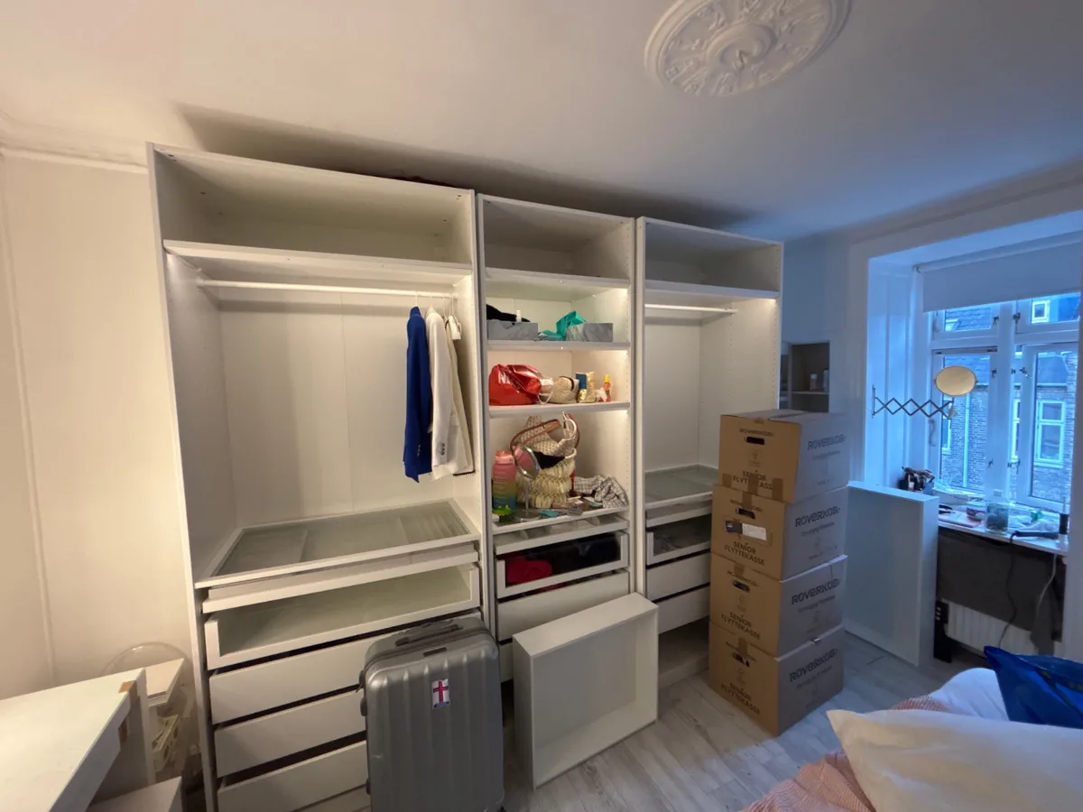 Et ikea pax skab med tøj, flyttekasser og en kuffert i et lyst værelse.