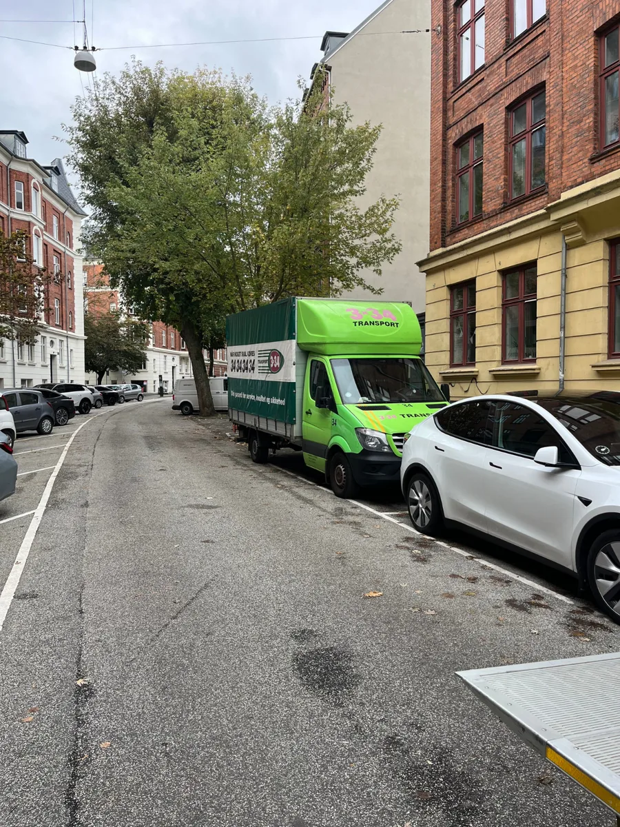 En grøn transportbil holder parkeret på en smal bygade mellem boligbygninger.